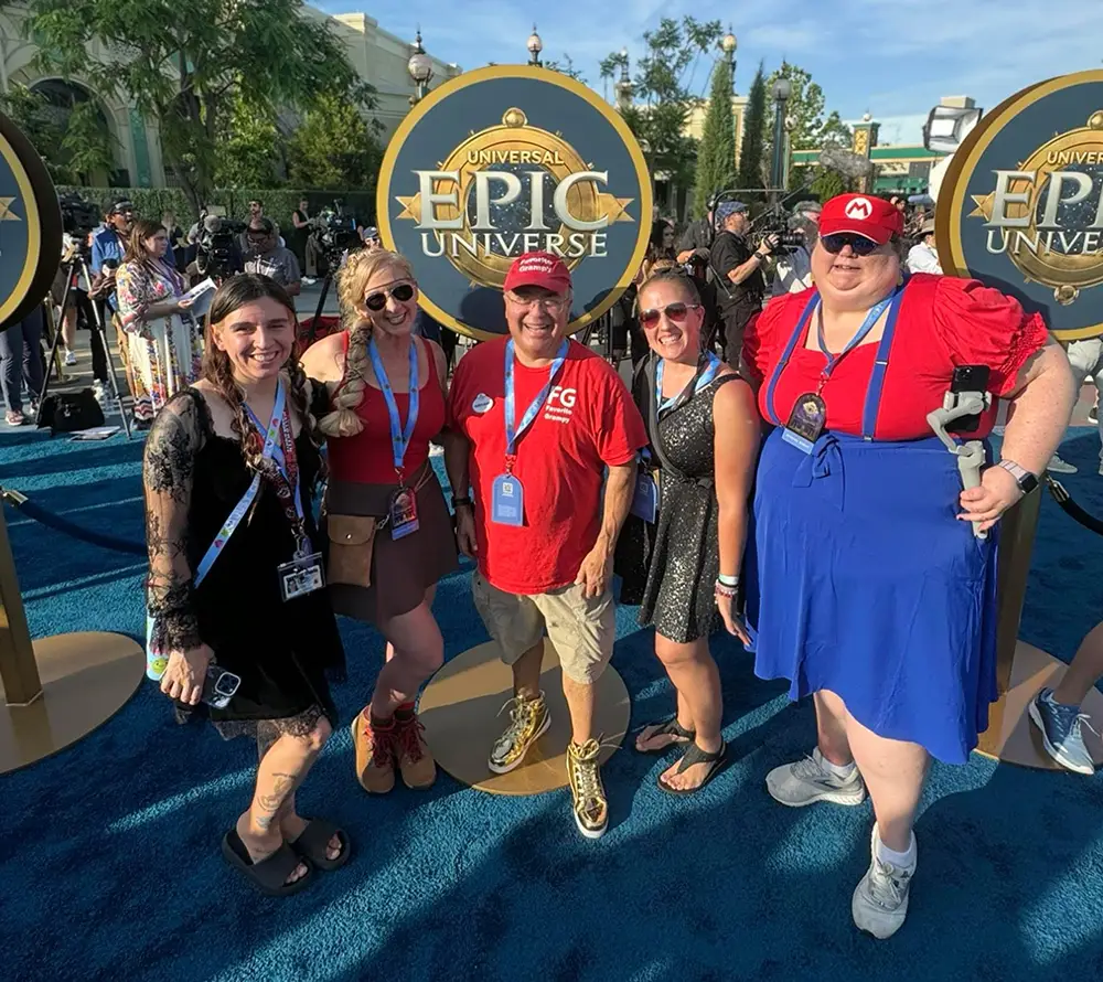 Five Smiling People Pose Together Outdoors at the Epic Universe Event, Standing on Blue Carpet Near Event Signs. Some Wear Red and Blue Outfits, Badges, and Lanyards, Enjoying a Sunny, Festive Atmosphere During Their Epic Universe Visit. - Favorite Grampy Travels - Favorite Grampy Travels 2026