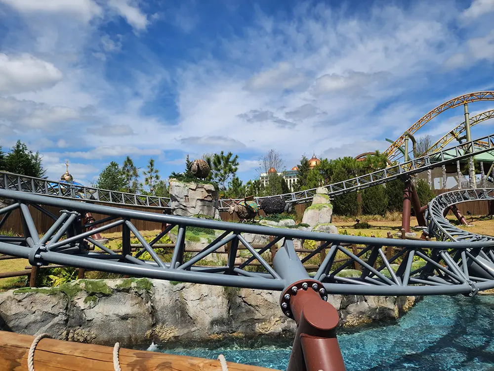 a Roller Coaster with Twisting Metal Tracks Set Above Rocky Terrain and Turquoise Water, Surrounded by Greenery and Buildings, Under a Partly Cloudy Blue Sky. - Favorite Grampy Travels - Favorite Grampy Travels 2026