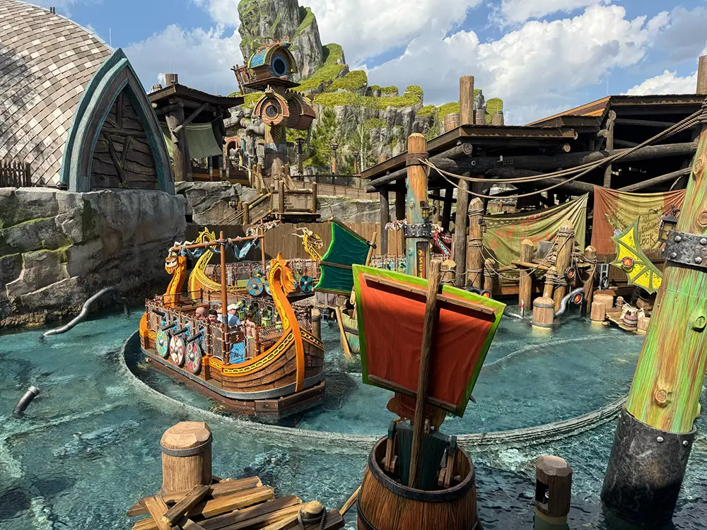 Uor-epic-universe-httyd-fyre-drill-overview - Favorite Grampy Travels a Colorful Viking-themed Boat Ride Splashes Through Water in an Amusement Park, Surrounded by Green Hills, Docks, and Rustic Wooden Scenery Under a Partly Cloudy Sky. - Favorite Grampy Travels - Favorite Grampy Travels 2026