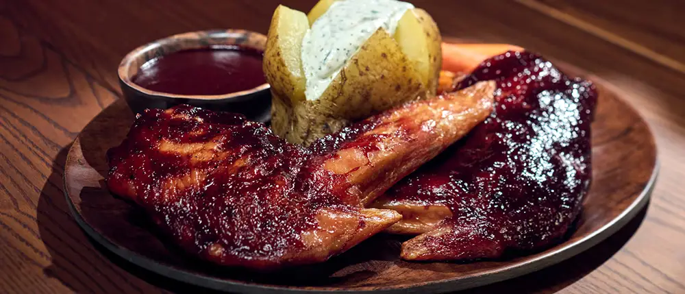 a Wooden Plate with Glazed Barbecue Ribs, a Baked Potato Topped with Sour Cream and Herbs, a Bowl of Barbecue Sauce, and Carrot Sticks on a Wooden Table—perfect for Fans Exploring Epic Universe Dining or Isle of Berk Restaurants. - Favorite Grampy Travels - Favorite Grampy Travels 2026