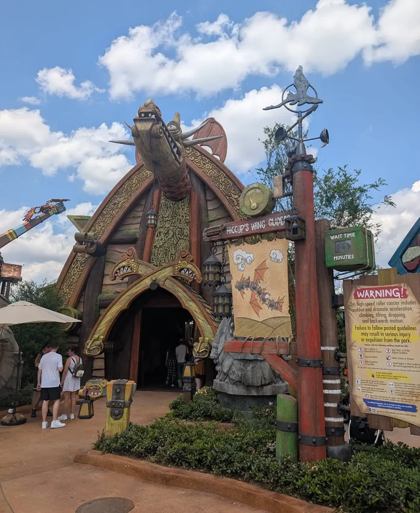 Uor-epic-universe-httyd-hiccups-wing-gliders-entrance - Favorite Grampy Travels the Entrance to a Theme Park Attraction Called Hiccup’s Wing Gliders, Featuring a Large Dragon Head Above the Doorway, Colorful Signs, and Guests Standing Nearby Under a Partly Cloudy Sky. - Favorite Grampy Travels - Favorite Grampy Travels 2026