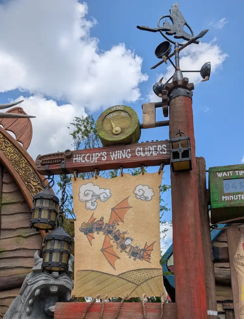 a Whimsical Sign for Hiccups Wing Gliders Features a Dragon and People Riding It, with Decorative Lanterns, a Weathervane, and a Partly Cloudy Sky in the Background. - Favorite Grampy Travels - Favorite Grampy Travels 2026