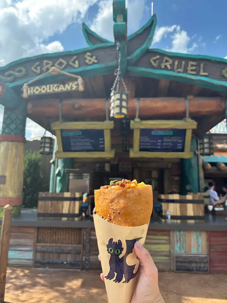 a Hand Holds a Savory Crepe Wrapped in Paper with a Cartoon Dragon Near the “grog & Gruel” Food Stand at Epic Universe’s Isle of Berk, Beneath a Blue Sky. Menu Boards Highlight Unique Dining Options Amid Rustic Decor in the Background. - Favorite Grampy Travels - Favorite Grampy Travels 2026