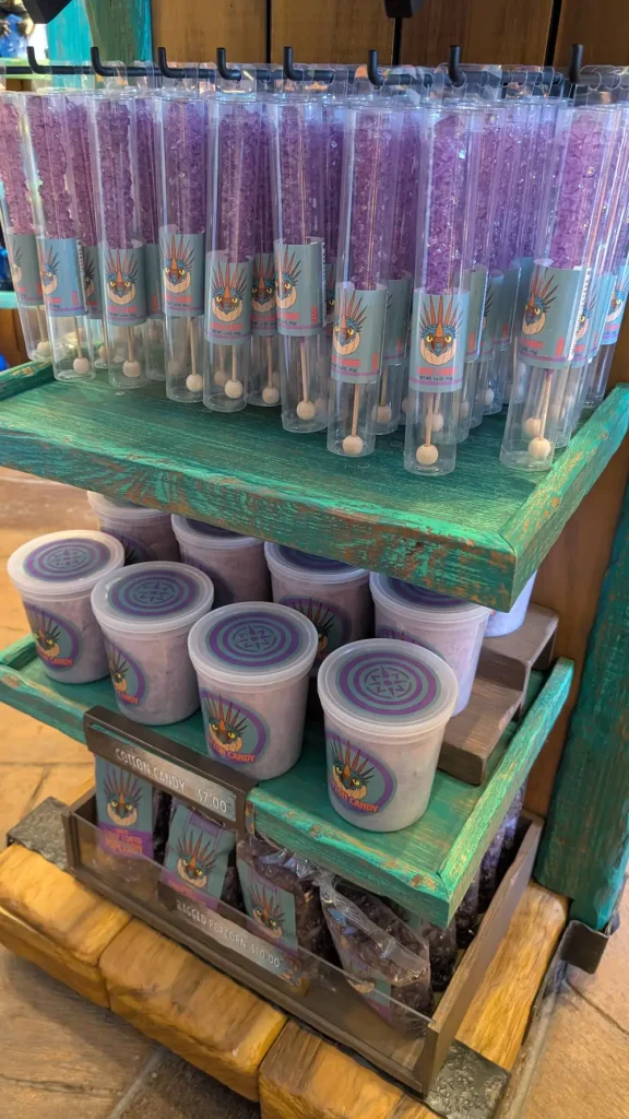 a Wooden Display at Epic Universe Features Clear Tubes of Purple Rock Candy and Round Containers of Purple Cotton Candy, All Adorned with Colorful Labels Showcasing a Stylized Bird Logo. Prices Are Visible for These Sweet Dining Options. - Favorite Grampy Travels - Favorite Grampy Travels 2026