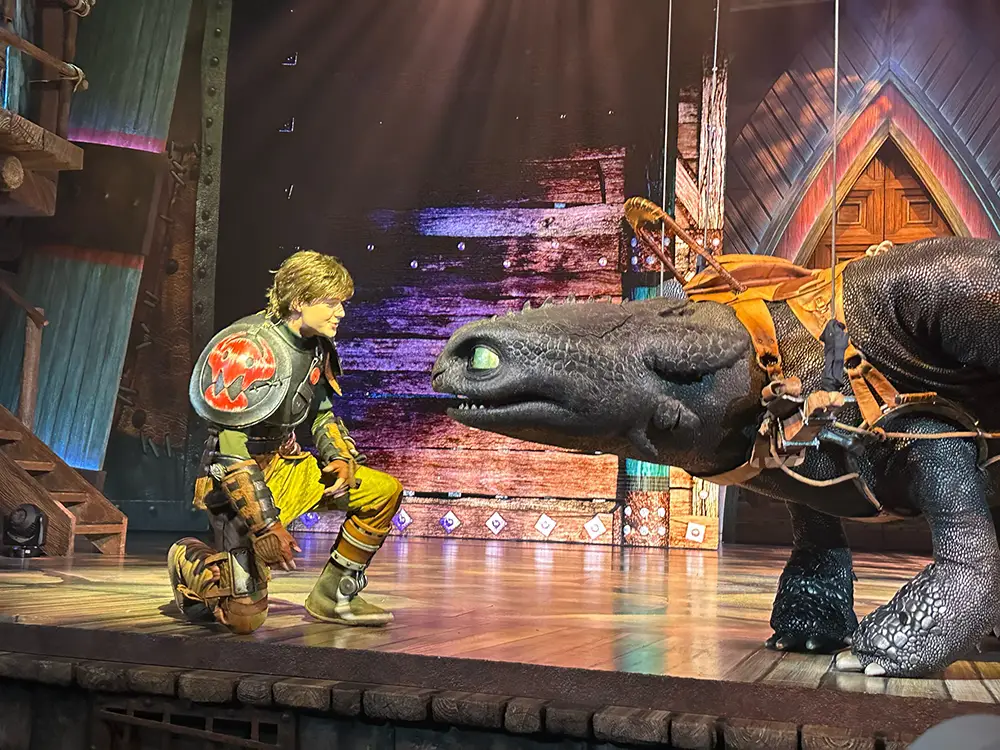 Uor-epic-universe-httyd-the-untrainable-dragon-toothless-hiccup-close-up - Favorite Grampy Travels a Performer Dressed As Hiccup Kneels and Faces a Large, Detailed Dragon Puppet Resembling Toothless on a Brightly Lit Stage with Wooden Scenery. - Favorite Grampy Travels - Favorite Grampy Travels 2026