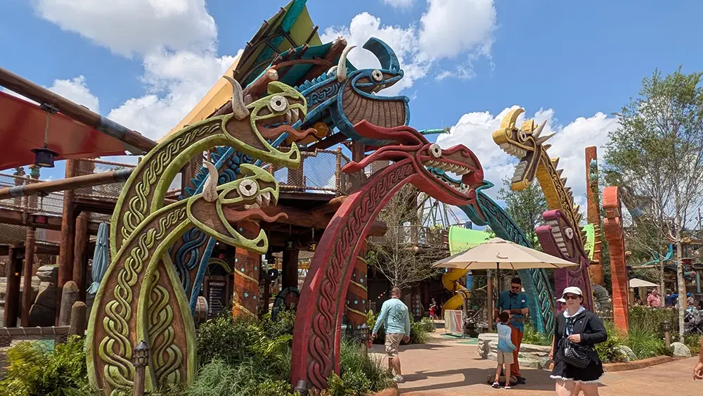 People Walk Through a Theme Park Entrance Decorated with Large, Colorful Norse Dragon Heads and Viking Ship Motifs, Inspired by the Isle of Berk, Under a Bright Blue Sky. Trees and Greenery Surround the Epic Universe Attractions. - Favorite Grampy Travels - Favorite Grampy Travels 2026