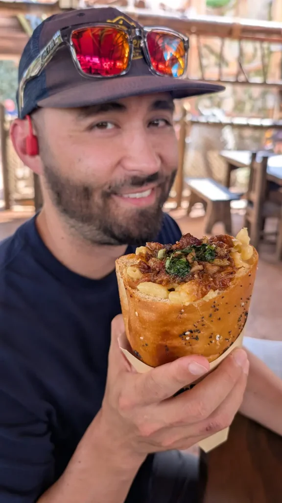 a Man with a Beard and Sunglasses on His Cap Smiles While Holding a Large Bread Cone Filled with Macaroni and Cheese, Bacon, and Herbs at an Outdoor Restaurant in Universal’s Epic Universe—perfect for a Tasty Family Outing. - Favorite Grampy Travels - Favorite Grampy Travels 2026