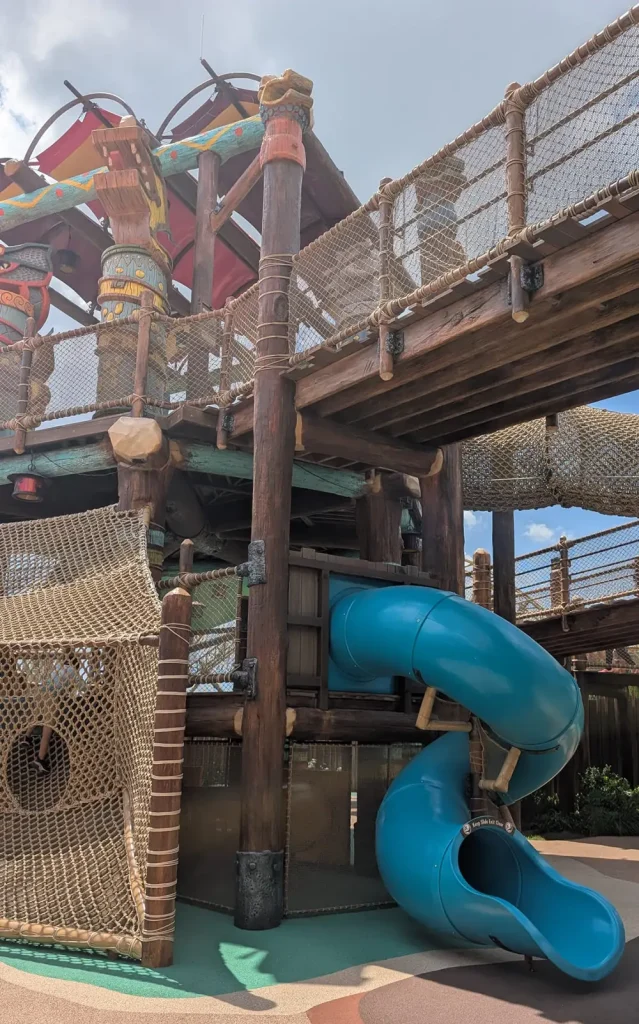 a Wooden Play Structure for Kids with Rope Bridges, Netting, and a Curved Blue Tube Slide. the Multi-level Setup is Outdoors Under a Partly Cloudy Sky—perfect for Toddlers’ Adventures Worthy of an Epic Universe. - Favorite Grampy Travels - Favorite Grampy Travels 2026