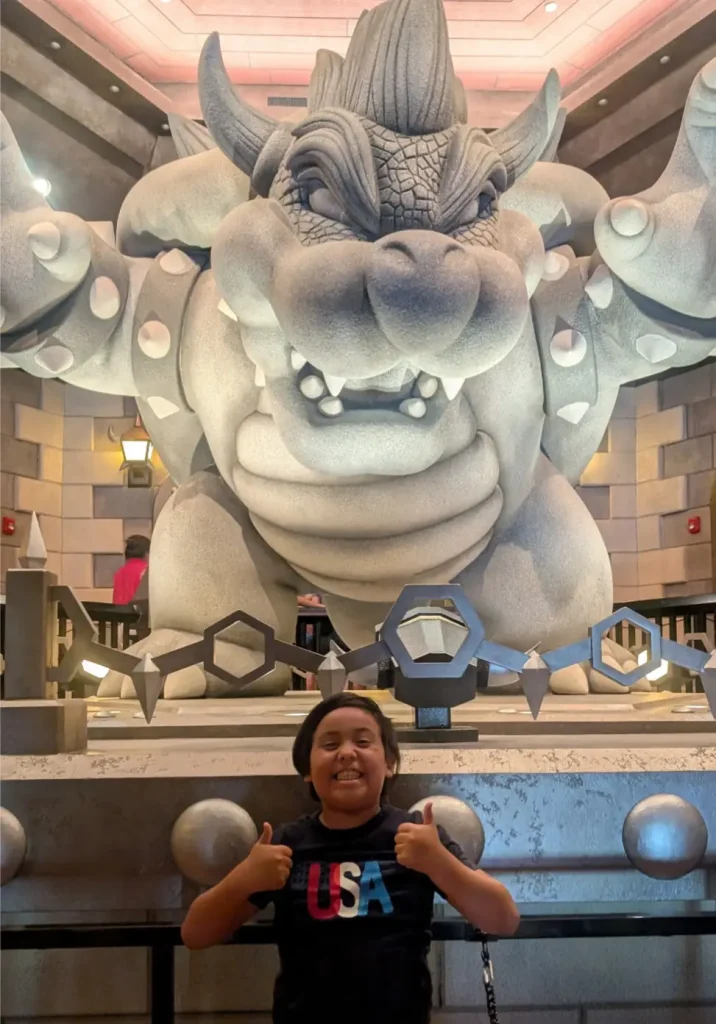 A smiling child wearing a black USA shirt gives two thumbs up in front of a large stone Bowser statue at a themed attraction, making it a perfect family photo spot for toddlers and kids alike. - Favorite Grampy Travels