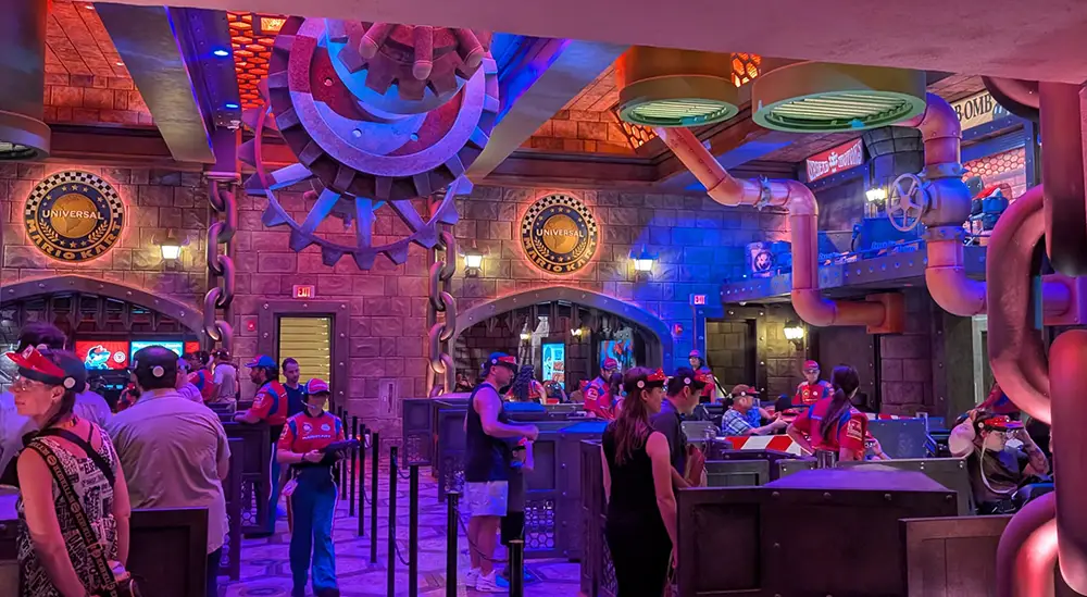 Families and Kids Wait in Line Inside a Dimly Lit, Steampunk-themed Universal Amusement Park Attraction with Gears, Pipes, and Universal Symbols on the Walls. Staff Members in Red Shirts Assist Guests Wearing Hats and Theme Park Gear. - Favorite Grampy Travels - Favorite Grampy Travels 2026