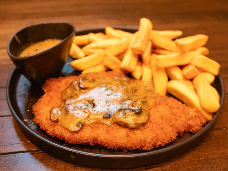 a Black Plate with a Breaded Schnitzel Topped with Mushroom Gravy, Thick Golden Fries, and a Small Black Bowl of Extra Sauce Awaits, Making Dining Here Feel Like a Magical Feast Straight from the Ministry of Magic on a Wooden Surface. - Favorite Grampy Travels - Favorite Grampy Travels 2026