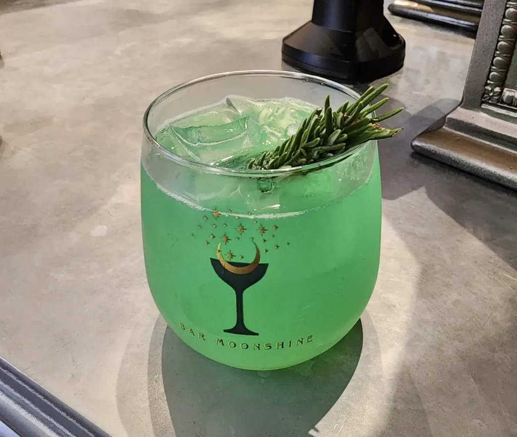 a Glass of Bright Green Cocktail with Ice Cubes and a Sprig of Rosemary Garnish, Served in a Round Glass Labeled Bar Moonshine, Awaits on a Gray Countertop—perfect for Magical Dining Worthy of the Ministry of Magic. - Favorite Grampy Travels - Favorite Grampy Travels 2026
