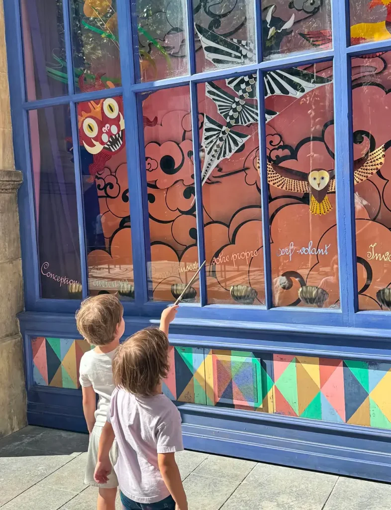 Two young children stand in front of a colorful shop window decorated with whimsical owl and bird displays, reminiscent of the Ministry of Magic at Epic Universe. One child points excitedly at the window with a wand as sunlight casts gentle shadows on the scene. - Favorite Grampy Travels