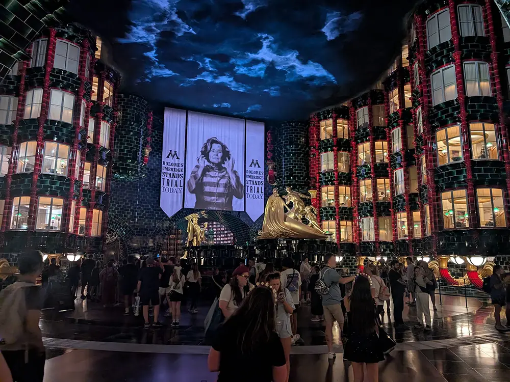 Nighttime View of a Circular Atrium with Glass-walled Buildings, Banners Reading 'dolores Umbridge Stands Trial Today,' and a Large Golden Statue Among a Crowd of Visitors. - Favorite Grampy Travels 2026