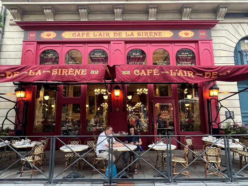 A Parisian café with red awnings and ornate signage reading “Café L’Air de la Sirène.” Two people sit outside, surrounded by empty wicker chairs and small round tables—like a scene from the magical world of Harry Potter. - Favorite Grampy Travels