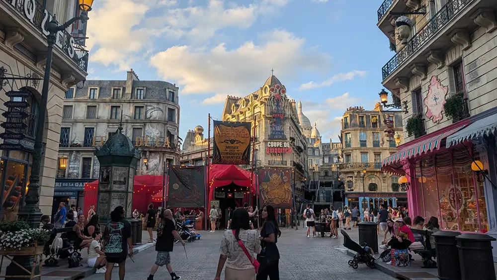 Busy pedestrian street in a theme-park style plaza with red market tents, ornate European buildings, and people strolling under a blue sky.