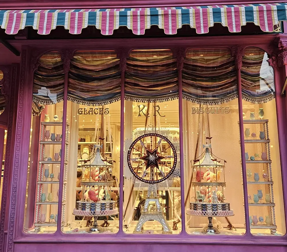 a Decorated Shop Window with Pink Trim Displays Colorful Pastries, a Miniature Eiffel Tower, and Ornate Carnival-themed Decorations—evoking the Magic of the Ministry of Magic. Striped Awnings and Gold Curtains Frame “glaces” and “biscuits” on the Glass. - Favorite Grampy Travels - Favorite Grampy Travels 2026