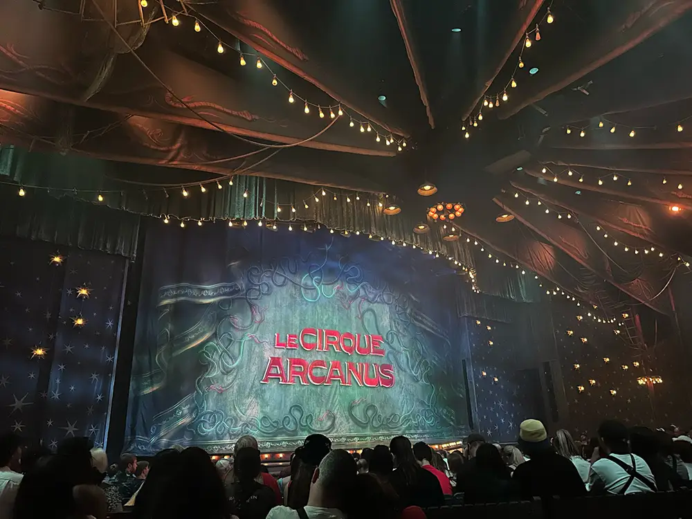 a Crowd Sits Facing a Stage with a Blue Curtain Displaying Le Cirque Arcanus in Red Letters. Strings of Lights Hang from the Ceiling, Evoking the Magical World of Harry Potter at Epic Universe. - Favorite Grampy Travels - Favorite Grampy Travels 2026