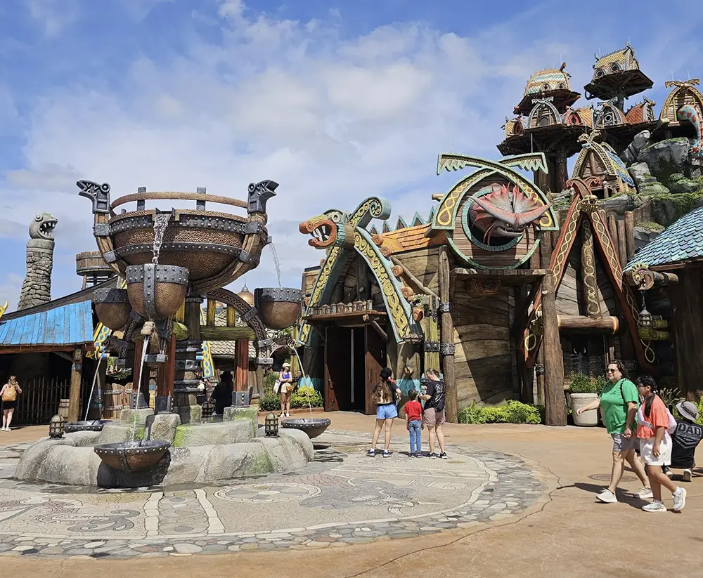 Uor-epic-universe-httyd-village-berk-fountain - Favorite Grampy Travels Visitors Walk Around a Viking-themed Attraction with Large Dragon Heads, Wooden Buildings, and Decorative Sculptures Under a Partly Cloudy Sky at an Amusement Park. - Favorite Grampy Travels - Favorite Grampy Travels 2026
