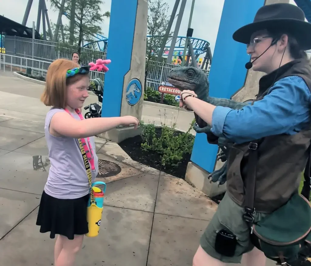 a Young Girl Smiles and Holds out Her Arm Toward a Person in a Ranger Outfit Holding a Realistic Dinosaur Puppet—a Vip Moment at Universal Kids Resort. Theme Park Rides and Another Smiling Person Are Visible in the Background. - Favorite Grampy Travels - Favorite Grampy Travels 2026