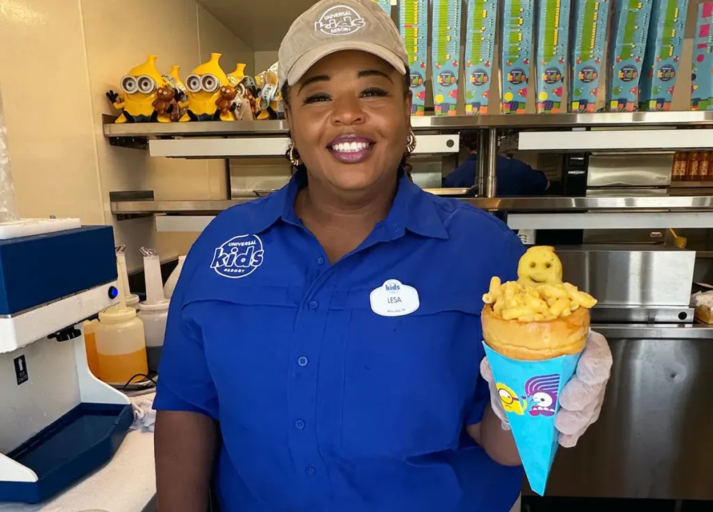 a Smiling Woman in a Blue Uniform and Tan Cap Holds a Cone of French Fries and a Potato Smiley Face, Standing in a Food Service Area Decorated with Minion Toys—perfect Snack for Guests Enjoying the Universal Kids Resort Vip Tour. - Favorite Grampy Travels - Favorite Grampy Travels 2026
