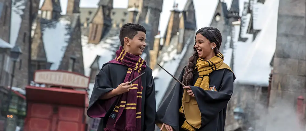 Two Kids in Wizard Robes and Scarves, Holding Wands and Smiling at Each Other, Stand in a Snowy, Magical Village Setting with Old-fashioned Buildings in the Background. - Favorite Grampy Travels - Favorite Grampy Travels 2026