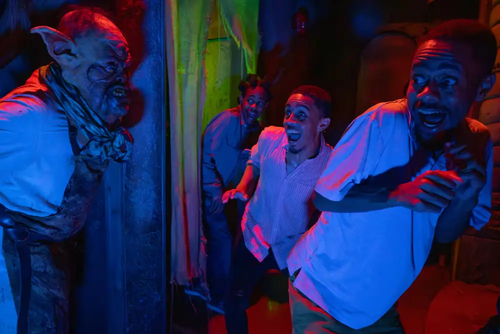 Three People Recoil in Fear As a Menacing Goblin-like Creature Confronts Them in a Dimly Lit House, Reminiscent of the Chilling Experiences at Universal Orlando's Halloween Horror Nights, with Dramatic Blue and Green Lighting. - Favorite Grampy Travels - Favorite Grampy Travels 2026