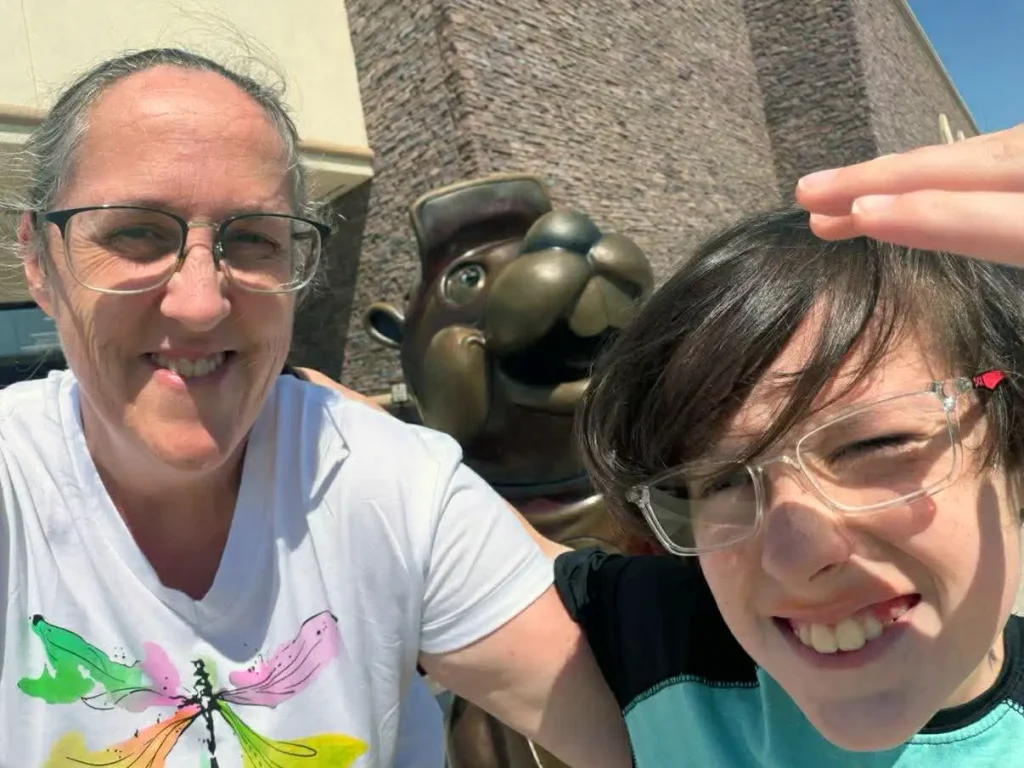 a Smiling Woman and a Child Pose for a Selfie Outside, with a Bucees Statue Behind Them. the Woman Wears Glasses and a Shirt with a Dragonfly, While the Child Squints and Shields Their Eyes from the Texas Sun. - Favorite Grampy Travels - Favorite Grampy Travels 2026