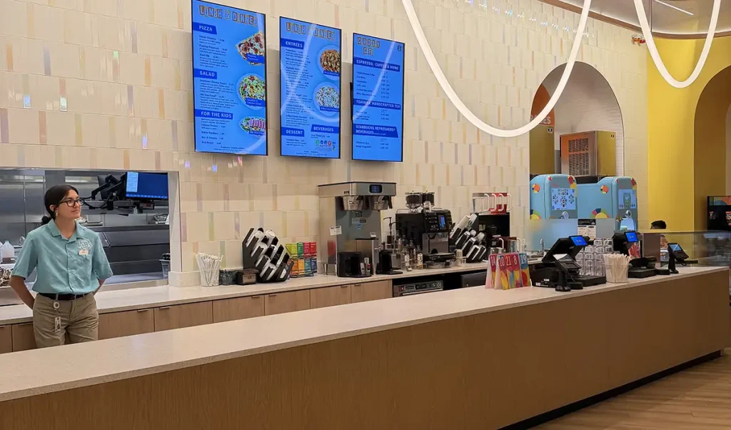 a Woman in a Teal Uniform Stands Behind a Modern, Brightly Lit Counter at Curiosity Cafe at Universal Kids Resort Hotel, with Digital Menu Boards, Coffee Machines, and Colorful Decor Visible in the Background. - Favorite Grampy Travels - Favorite Grampy Travels 2026