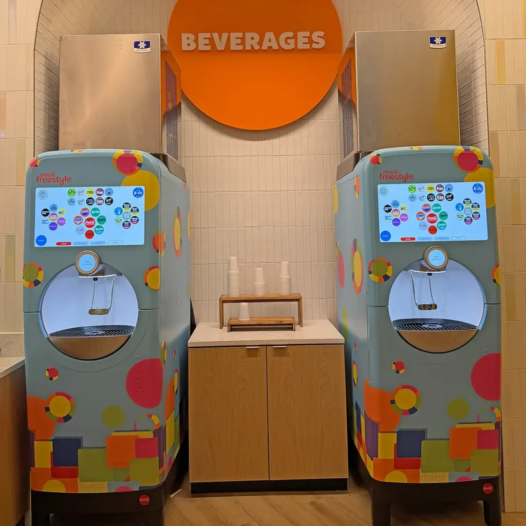 Two Modern Soda Fountain Machines with Colorful Designs Stand Side by Side, with a Wooden Counter and Stack of Plastic Cups—including Freestyle Refillable Cups at Universal Kids Resort Hotel—in Between. a Large Orange “beverages” Sign is Displayed Above Them. - Favorite Grampy Travels - Favorite Grampy Travels 2026