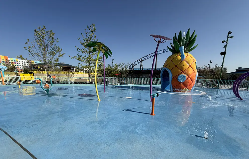 Children’s Splash Pad with Colorful Water Features, Including a Large Pineapple Structure and Palm Tree Sprayers, Offers the Perfect Setting for Universal Kids Resort Rides Excitement on Your First Day, All Under a Sunny Sky with Nearby Buildings in the Background. - Favorite Grampy Travels - Favorite Grampy Travels 2026