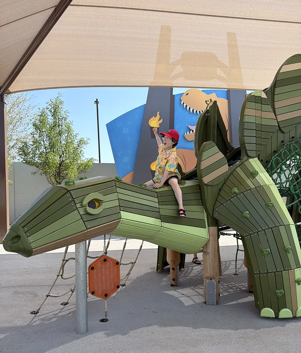 a Child in a Red Cap and Colorful Shirt Sits Atop a Green Dinosaur Playground Structure, Reaching Up with One Hand. a Shaded Canopy and a Dinosaur Mural Are Visible in the Background. - Favorite Grampy Travels - Favorite Grampy Travels 2026