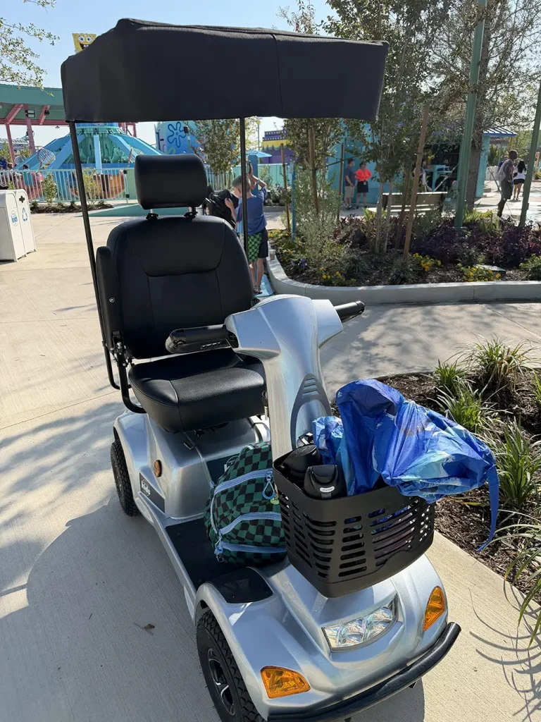 a Silver Mobility Scooter with a Black Canopy is Parked Outdoors, Perhaps Ready for Its First Day Exploring Universal Kids Resort Rides. a Green Plaid Bag and a Blue Poncho Are in Its Front Basket. Sunlit Pathways, Greenery, and People Are Visible in the Background. - Favorite Grampy Travels - Favorite Grampy Travels 2026