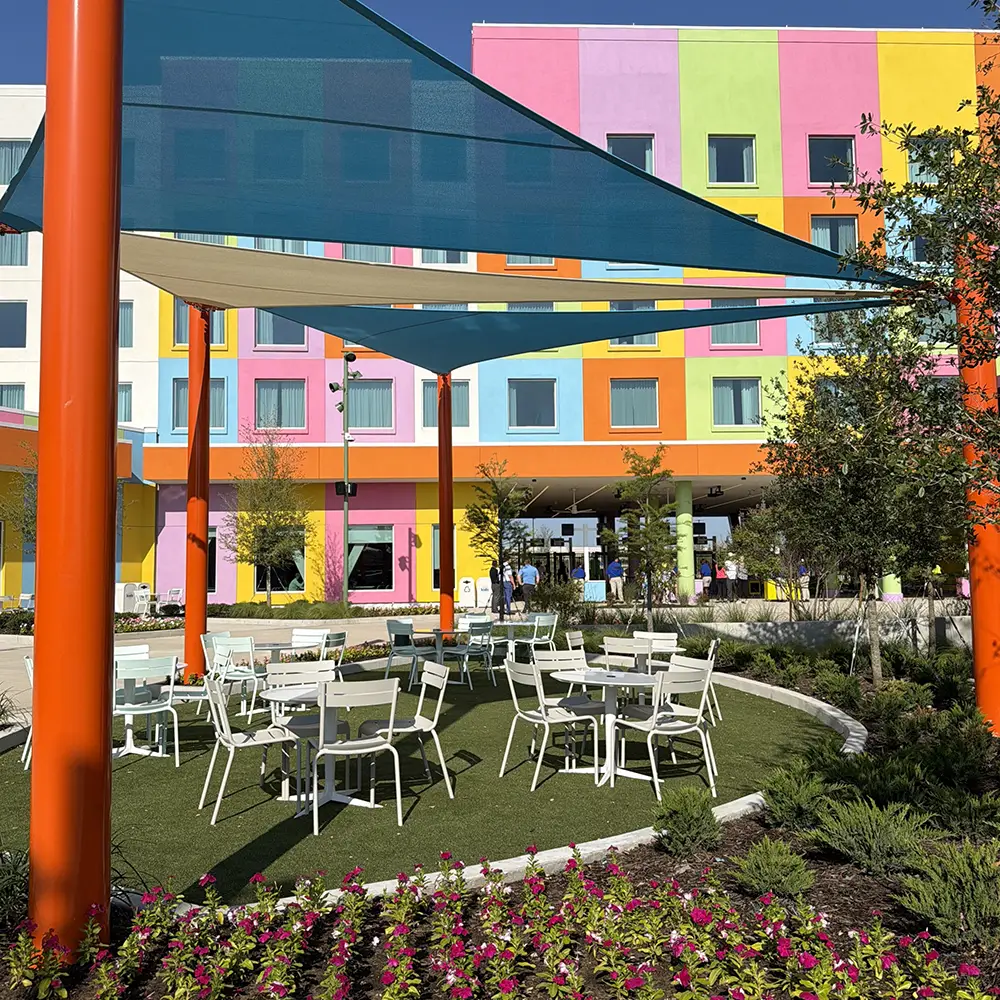 Colorful Building with Square Windows in the Background, Reminiscent of a Universal Kids Resort Rides Area, Outdoor Seating Area with White Tables and Chairs, Blue Shade Sails Overhead, Vibrant Flowers, and Greenery in the Foreground—creating a Lively Atmosphere Perfect for Enjoying Your First Day. - Favorite Grampy Travels - Favorite Grampy Travels 2026