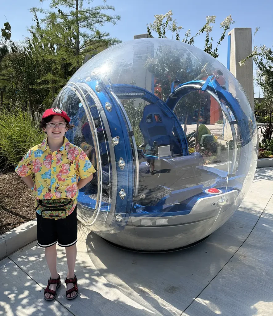 a Smiling Child in a Colorful Shirt and Red Cap Stands Next to a Large, Transparent Spherical Vehicle with Blue Seats, Outdoors on a Sunny Day—ready for Their First Day Experiencing Universal Kids Resort Rides. - Favorite Grampy Travels - Favorite Grampy Travels 2026