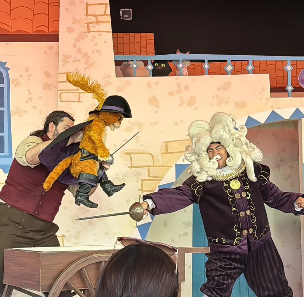 a Performer Holds a Puppet of a Cat Wearing a Hat and Cape, While Another Actor in a White Wig and Ornate Costume Wields a Sword on Stage. the Scene Appears Lively and Theatrical, Reminiscent of the Excitement Children Feel on Their First Day Exploring Universal Kids Resort Rides, All Set Against a Colorful Backdrop. - Favorite Grampy Travels - Favorite Grampy Travels 2026