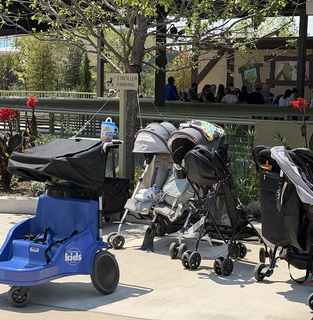 Several Strollers and a Blue Kids’ Cart Are Parked in a Designated Stroller Parking Area Outdoors Near a Shaded Pavilion, Suggesting That Families Are Taking a Break After Enjoying Universal Kids Resort Rides, Possibly on Their First Day at the Park. Trees and Plants Create a Pleasant Backdrop for This Family-friendly Scene. - Favorite Grampy Travels - Favorite Grampy Travels 2026