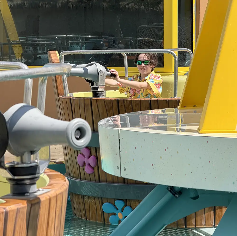 on Their First Day at Universal Kids Resort Rides, a Person Wearing Green Sunglasses Sits in a Water Ride Shaped Like a Wooden Barrel, Aiming a Water Cannon. Bright Decorations and Splashing Water Are Visible Around the Ride. - Favorite Grampy Travels - Favorite Grampy Travels 2026