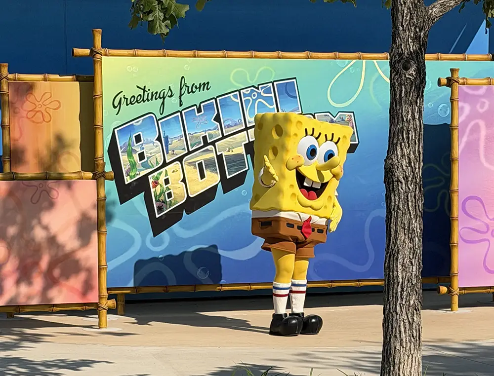 a Person in a Spongebob Squarepants Costume Stands in Front of a Colorful Greetings from Bikini Bottom Sign, Celebrating Their First Day at Universal Kids Resort Rides, with a Tree Partially Visible on the Right Side of the Image. - Favorite Grampy Travels - Favorite Grampy Travels 2026