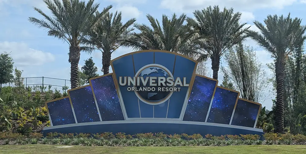 Uor-entrance-sign-driving - Favorite Grampy Travels a Large Universal Orlando Resort Sign with a Globe Logo Stands in Front of Tall Palm Trees and Lush Landscaping, Inviting Guests to Start Their Adventure with a Universal Orlando Vacation Package Under a Partly Cloudy Sky. - Favorite Grampy Travels - Favorite Grampy Travels 2026