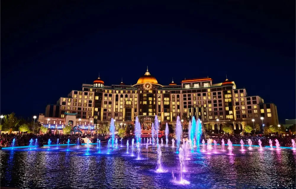 Uor-epic-universe-helios-grand-hotel-night-time-fountain - Favorite Grampy Travels a Large, Grand Building at Universal Orlando is Illuminated at Night with Colorful Lights. in Front, a Fountain Displays Vibrant Streams of Water in Blue, Pink, and Purple Hues, Drawing Vacation Package Guests to Watch the Dazzling Show. - Favorite Grampy Travels - Favorite Grampy Travels 2026