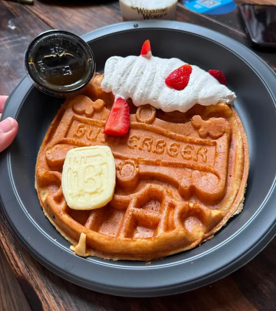 a Round Waffle Shaped Like the Hogwarts Express, Topped with a Pat of Butter, Strawberries, Whipped Cream, and Served with a Small Container of Syrup on a Black Plate. - Favorite Grampy Travels - Favorite Grampy Travels 2026