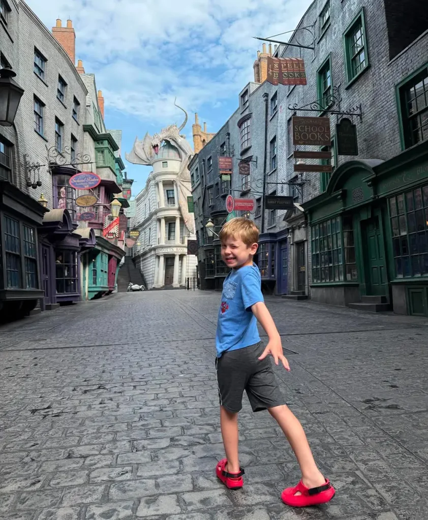 a Young Boy in a Blue Shirt and Red Shoes Excitedly Poses on a Cobblestone Street in a Harry Potter-themed Area, with a Dragon-topped Building in the Background Under a Blue Sky. - Favorite Grampy Travels - Favorite Grampy Travels 2026