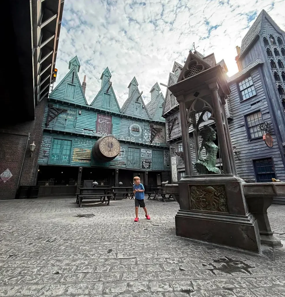 a Young Boy Stands in a Cobblestone Courtyard with Whimsical, Pointed-roof Buildings and a Statue Fountain, Under a Partly Cloudy Sky. the Scene Has a Magical, Fantasy Atmosphere. - Favorite Grampy Travels - Favorite Grampy Travels 2026