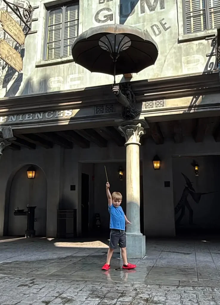 a Young Boy Stands on a Cobblestone Street, Holding a Wand Up Toward a Large Umbrella Attached to a Building Facade with Whimsical Signage and Shadowy Figures on the Wall Behind Him. - Favorite Grampy Travels - Favorite Grampy Travels 2026