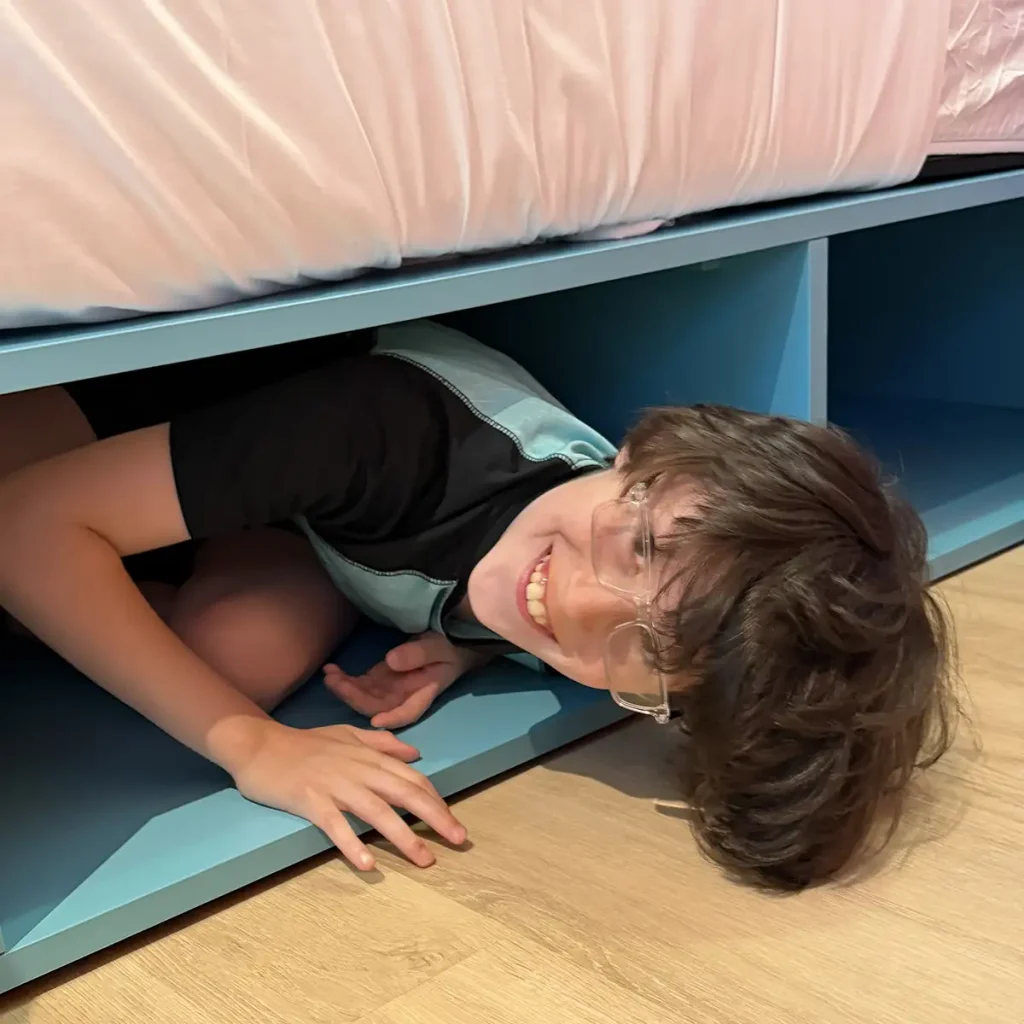 a Smiling Child Wearing Glasses and a Black Shirt is Playfully Crawling Under a Blue Bed Frame, Playing Hid N Seek in His Hotel Room at Universal Kids Resort, Looking Up at the Camera. - Favorite Grampy Travels - Favorite Grampy Travels 2026