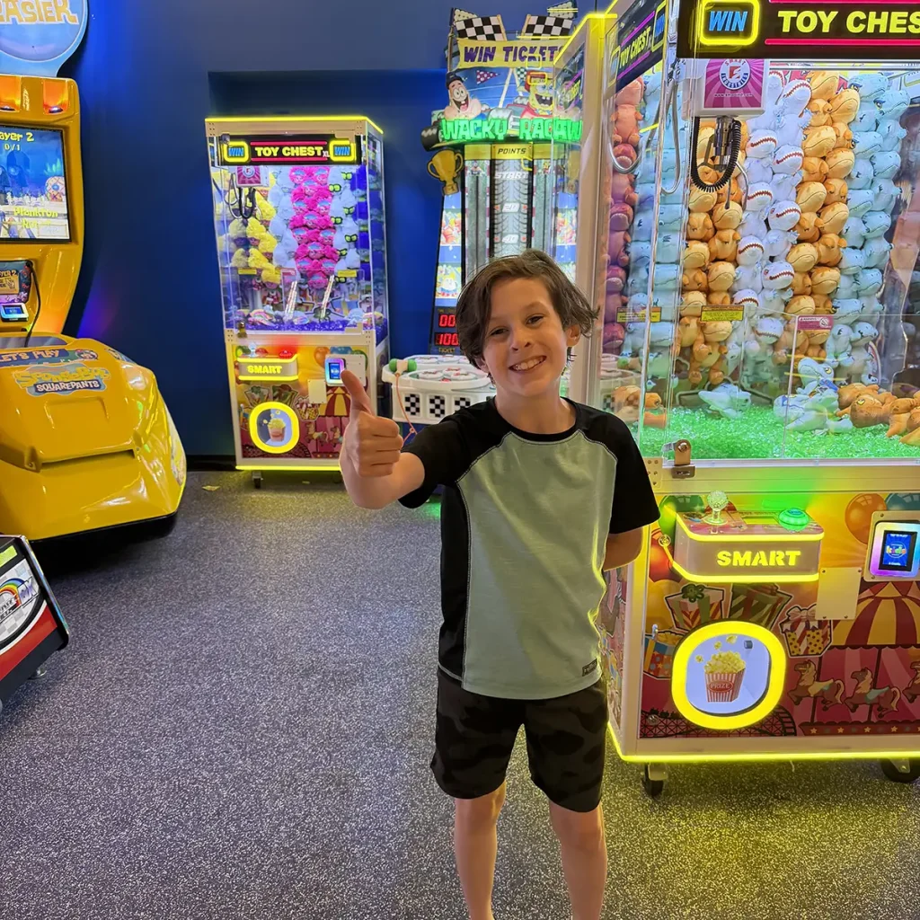 a Smiling Boy in a Light Green and Black Shirt Gives a Thumbs Up While Standing in Front of Arcade Claw Machines and Games, with Bright Lights in the Background. - Favorite Grampy Travels - Favorite Grampy Travels 2026