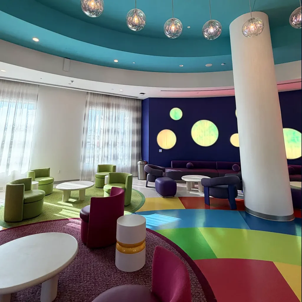 a Colorful Modern Lounge, Reminiscent of the Universal Kids Resort Hotel Lobby, with Round Chairs, Circular Tables, a Tall White Column, Vibrant Flooring, and Wall Lights Shaped Like Glowing Dots. Large Windows with Sheer Curtains Let in Natural Light. - Favorite Grampy Travels - Favorite Grampy Travels 2026