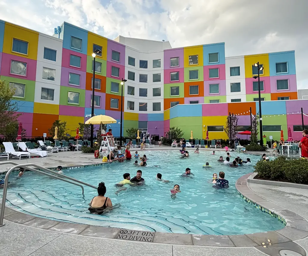 People Swim and Relax in the Universal Kids Resort Hotel Pool, Surrounded by Colorful, Geometric-patterned Walls in the Background. Some Sit on Lounge Chairs While Others Play in the Water Under a Cloudy Sky. - Favorite Grampy Travels - Favorite Grampy Travels 2026