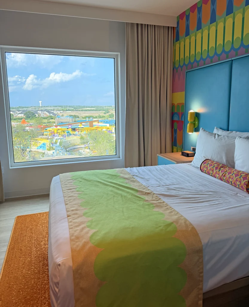a Brightly Decorated Hotel Room at Universal Kids Resort in Fisco, Texas, with a Neatly Made Bed and a Large Window Showing a View of a Colorful Amusement Park and Blue Sky Outside. - Favorite Grampy Travels - Favorite Grampy Travels 2026